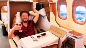 Pasajero sonriente relajado en asiento privado de clase ejecutiva de avión, recostado con almohada y copa de champán junto a una bandeja con postre y cubiertos, interiores de cabina con acabados de madera y ventanillas al cielo, Gds global distribution system ventajas y desventajas esenciales.