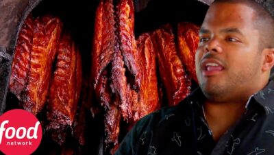 Persona sin identificar hablando frente a costillas ahumadas colgando en el interior de un ahumador con brillo caramelizado y textura jugosa, imagen que sugiere una explicación sobre técnicas culinarias, sabor, seguridad alimentaria y conservación, Ventajas y desventajas del ahumado de alimentos: secretos y riesgos