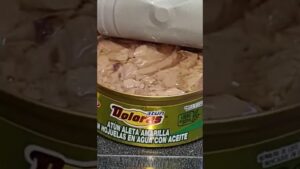 Lata de atún abierta en primer plano con trozos rosados sumergidos en líquido y tapa parcialmente levantada, etiqueta verde que muestra "atún aleta amarilla en hojuelas en agua con aceite" y textura visible de las fibras del pescado, Ventajas y desventajas del atún enlatado: ¿es saludable para ti?