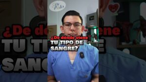 Hombre joven con gafas y uniforme médico azul mirando a cámara en un video vertical con texto superpuesto sobre el origen del tipo de sangre, placa de reconocimiento y modelos anatómicos en el fondo, optimizado para búsquedas sobre Sangre B positivo ventajas y desventajas que debes conocer