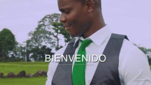 Hombre sonriente con camisa blanca, corbata verde y chaleco negro mirando hacia abajo en un paisaje de césped y árboles con la palabra BIENVENIDO superpuesta en el centro, Banca premier Caixabank ventajas y desventajas: ¿vale la pena?