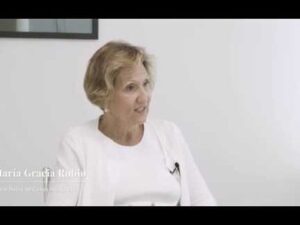 Mujer adulta sentada en una sala blanca, vestida con chaqueta y camiseta blancas y con micrófono de solapa mientras participa en una entrevista de formato profesional, imagen nítida y minimalista que ilustra Ventajas y desventajas de la banca privada en pagos sociales.
