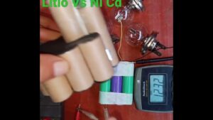 Fotografía de una demostración práctica con manos sosteniendo un paquete de celdas cilíndricas, un pequeño conjunto de baterías envueltas y cableadas, bombillas incandescentes, pinzas y un multímetro sobre una mesa que ilustra una comparativa y pruebas rápidas, Baterías ni cd ventajas y desventajas: rendimiento y usos clave