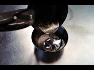 Primer plano de metal fundido brillante siendo vertido desde un cucharón sujetado con alicates hacia un pequeño recipiente metálico sobre una superficie de acero, con reflejos, sombras y texturas que comunican procesos de fundición, seguridad y reciclaje, útil como imagen ilustrativa para Baterías de zinc ventajas y desventajas: impacto real en energía