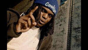 Persona joven con gorro azul con logo, rastas y chaqueta oscura señalando con el dedo índice frente a una pared metálica desgastada, con luz lateral cálida que realza texturas y una expresión seria y directa; Ventajas y desventajas de Be Real: descubre si esta app es para ti