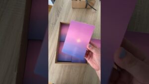 Una mano con las uñas pintadas sostiene una tarjeta o libreta con un degradado rosa a azul y un sol tenue, sobre una caja con varias unidades idénticas en una mesa de madera — Bloc de notas ventajas y desventajas: ¿vale la pena usarlo?