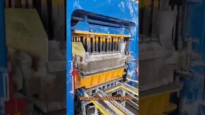 Máquina industrial azul y amarilla que moldea bloques de hormigón con moldes metálicos y cinta transportadora, con restos de cemento visibles, ideal para ilustrar contenido técnico y accesible sobre Bloque de hormigón ventajas y desventajas para tu construcción.