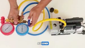 Vista cenital de manos de técnico ajustando un juego de manómetros con carátulas roja y azul y mangueras amarilla y azul conectadas a una bomba de vacío metálica sobre fondo blanco, mostrando válvulas, acoples y accesorios para mantenimiento HVAC y seguridad, orientada a quienes comparan equipos y necesitan información práctica, Ventajas y desventajas bomba de vacío: guía esencial para elegir bien.