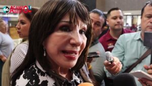 Mujer en primer plano con cabello castaño oscuro y maquillaje pronunciado hablando ante varios micrófonos y reporteros en un espacio interior, con teléfonos y rostros de fondo, imagen clara y detallada para acompañar un artículo o reportaje sobre Ventajas y desventajas del crecimiento de plantas en invernadero y exterior.