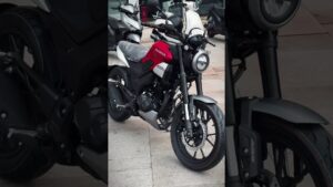 Honda CB 190 ventajas y desventajas: descubre si esta moto es para ti — vista frontal en tres cuartos de una moto naked roja y negra en un concesionario, con faro redondo LED, horquilla telescópica, freno de disco delantero, llantas de aleación, asiento cubierto con plástico protector y motor y escape visibles junto a scooters en segundo plano.