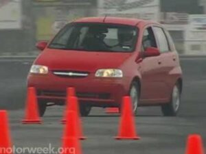 Compacto Chevrolet Aveo rojo captado en un ángulo frontal mientras maniobra entre conos naranjas en una pista de prácticas con conductor visible, texto accesible y optimizado para SEO sobre prueba de manejo y estabilidad, y Ventajas y desventajas del Chevrolet Aveo: ¿vale la pena comprarlo?