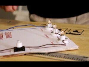 Placa de demostración sobre una mesa con varios portalámparas y bombillas alineadas conectadas por cables a un interruptor y manos al fondo manipulando el montaje — Ventajas y desventajas circuito en serie: descubre su impacto real