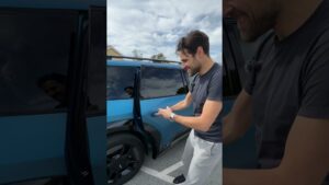 Hombre sonriente abriendo la puerta trasera de un SUV azul en un aparcamiento bajo un cielo nublado, con atención al diseño lateral y las llantas, evocando una comparación práctica sobre Ventajas y desventajas de un SUV para elegir tu coche ideal.