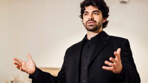 Orador vestido de negro gesticulando con las manos y mirando al frente sobre un fondo claro, transmitiendo seguridad y lenguaje corporal abierto, Ventajas y desventajas de comunicación oral: descubre su impacto real.