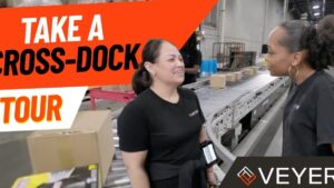 En un amplio almacén de cross docking, dos trabajadoras conversan junto a una cinta de rodillos con varias cajas mientras una sostiene un escáner de mano y un cartel naranja invita a una visita, Ventajas y desventajas de cross docking para tu logística