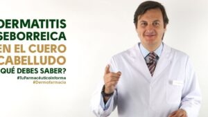 Hombre con bata blanca, corbata y placa identificativa sonriente señalando hacia la cámara frente a un fondo blanco con texto informativo sobre dermatitis seborreica en el cuero cabelludo, imagen clara y profesional pensada para ilustrar Ventajas y desventajas del romero en el cabello: ¿realmente funciona?