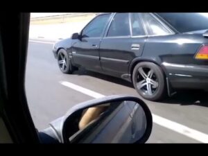 Fotografía tomada en movimiento desde el interior de un coche que muestra un Daewoo Espero negro de perfil con llantas deportivas de cinco radios y ventanas tintadas circulando por la carretera, reflejo del retrovisor y sensación de velocidad; Ventajas y desventajas del Daewoo Espero: ¿vale la pena comprarlo?