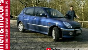 Vista lateral de un hatchback Daihatsu Sirion azul metálico aparcado sobre tierra en un claro boscoso, con las ruedas orientadas hacia la cámara, un presentador desenfocado detrás, una banda roja lateral con texto editorial y el logotipo de YouTube en la esquina, Ventajas y desventajas del Daihatsu Sirion: ¿vale la pena?