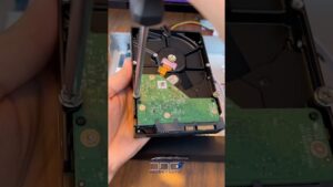 Mano que sostiene y desmonta un disco duro mecánico abierto con un destornillador aflojando tornillos sobre la placa de circuito verde y los conectores SATA visibles, escena de reparación o recuperación de datos que invita a analizar 'Ventajas y desventajas disco duro SSD: ¿vale la pena?' como comparativa entre HDD y SSD.