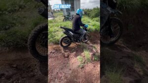 Persona con casco azul y guantes montada en una moto off-road negra sobre un camino de tierra y piedras junto a césped, con otra moto aparcada y un edificio al fondo, texto superpuesto que dice QUE FALLO? y Ventajas y desventajas de la dm 150: ¿vale la pena esta moto?