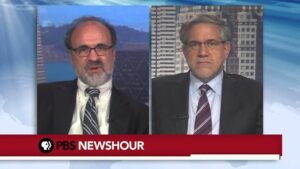 Pantalla dividida de un noticiero que muestra a dos hombres adultos con traje, corbata y gafas en una entrevista remota frente a fondos urbano y de estudio y la barra informativa de PBS NewsHour, contextualizando el tema Ventajas y desventajas del DSM V y su impacto en salud mental