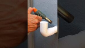 Imagen de una mano sujetando un soplete de mano con boquilla metálica que aplica calor a una curva de tubería blanca de PVC instalada en una pared gris, representando trabajo de fontanería y materiales, Qué ventajas y desventajas tiene el pvc: descubre si es ideal para ti