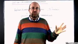 Hombre con gafas y jersey de rayas, de pie y gesticulando frente a una pizarra blanca con notas sobre empresas multinacionales, dando una explicación en clase o presentación sobre impactos económicos y sociales, Ventajas y desventajas de las multinacionales: impacto y claves