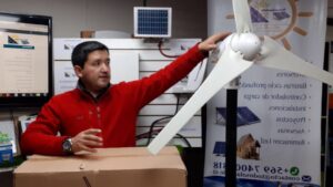 Un hombre con chaqueta roja muestra y señala las palas de una turbina eólica de pequeño tamaño montada en un soporte en un espacio interior con paneles solares y material informativo que sugiere una demostración didáctica, Ventajas y desventajas de la energía eólica: impacto real y futuro.