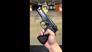 Mano sosteniendo una pistola Beretta 92 FS en un campo de tiro interior con dianas desenfocadas al fondo, con énfasis en el armazón, seguro y empuñadura texturizada para describir la escena, contexto de seguridad y uso responsable, Ventajas y desventajas de la F.S.N: descubre su impacto y retos clave