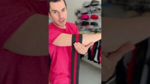 Hombre con camiseta roja en un espacio de accesorios deportivos hace una demostración sosteniendo una banda elástica negra con una franja roja enrollada alrededor del antebrazo frente a estantes con cinturones y bandas, Ventajas y desventajas de usar faja en el gym: ¿ayuda o perjudica?