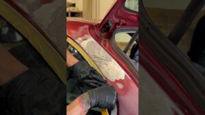Manos con guantes negros lijando y retirando pintura en la aleta de un auto rojo junto al marco del parabrisas, con cinta de enmascarar amarilla y chapa expuesta en un taller de carrocería, Ventajas y desventajas piletas de fibra de vidrio: ¿son para ti?