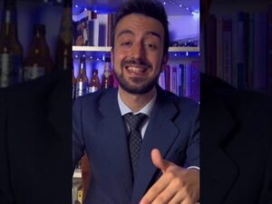 Hombre joven con traje y corbata sonriendo y gesticulando hacia la cámara en un video vertical, delante de una estantería con libros y botellas iluminadas por luces LED azules, como un asesor que ofrece una explicación clara y cercana sobre 'Ventajas y desventajas de fijo discontinuo: ¿conviene este contrato?'.