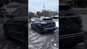 Ford Edge negro, vista trasera en tres cuartos, estacionado sobre asfalto con una ligera capa de nieve en techo y cristales, faros traseros y doble salida de escape visibles, llantas oscuras y entorno invernal con casas y árboles bajo cielo nublado, Ventajas y desventajas Ford Edge 2013: ¿vale la pena comprarlo?