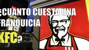 Imagen de un letrero luminoso con el rostro del Coronel Sanders y grandes letras sobre la fachada de un restaurante KFC al anochecer, con mesas exteriores visibles y un mensaje que pregunta por el costo de la franquicia, Ventajas y desventajas de la franquicia KFC: ¿es tu oportunidad ideal?
