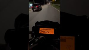 Vista en primera persona del tablero digital iluminado en naranja de una motocicleta circulando de noche por una vía urbana con un coche a la izquierda, transmitiendo sensación de alta velocidad y enfoque en el velocímetro, Yamaha FZ 250 ventajas y desventajas: descubre si esta moto es para ti.