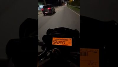 Vista en primera persona del tablero digital iluminado en naranja de una motocicleta circulando de noche por una vía urbana con un coche a la izquierda, transmitiendo sensación de alta velocidad y enfoque en el velocímetro, Yamaha FZ 250 ventajas y desventajas: descubre si esta moto es para ti.