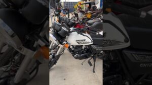 Vista angular de una motocicleta blanca Suzuki GN125F aparcada en un concesionario entre varias motos, con el logo y el depósito visibles, faro cromado, relojes gemelos en el manillar, intermitentes naranjas, motor monocilíndrico y pata de cabra desplegada, descripción accesible y optimizada para SEO que invita a analizar Ventajas y desventajas de Suzuki GN 125: ¿la moto ideal para ti?
