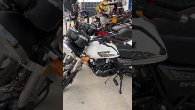 Vista angular de una motocicleta blanca Suzuki GN125F aparcada en un concesionario entre varias motos, con el logo y el depósito visibles, faro cromado, relojes gemelos en el manillar, intermitentes naranjas, motor monocilíndrico y pata de cabra desplegada, descripción accesible y optimizada para SEO que invita a analizar Ventajas y desventajas de Suzuki GN 125: ¿la moto ideal para ti?