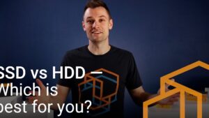 Presentador sonriente de pie ante un fondo azul muestra cajas y componentes de almacenamiento mientras en la imagen aparece texto superpuesto en blanco "SSD vs HDD" y elementos geométricos naranjas, Ventajas y desventajas HDD y SSD: descubre cuál es mejor para ti