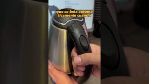 Primer plano de manos sujetando el asa negra de un hervidor eléctrico de acero inoxidable con el pulgar sobre el botón de apertura junto a la tapa y un fondo de escritorio desenfocado; Ventajas y desventajas del hervidor eléctrico: ¿vale la pena?