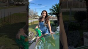 Joven sonriente con overol corto de mezclilla apoyada en el capó verde de un coche mientras sostiene un bidón de aceite de motor verde en un jardín residencial soleado con palmera y reflejo en la carrocería — Ventajas y desventajas del Hyundai Elantra 2013 para comprar