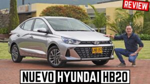 Fotografía frontal tres cuartos de un Hyundai HB20 plateado estacionado sobre un camino de adoquines en un jardín residencial, con un hombre agachado junto al paragolpes derecho presentando el vehículo y resaltando parrilla hexagonal, faros delanteros, llantas de aleación y silueta compacta, Ventajas y desventajas del Hyundai HB20 para elegir tu auto ideal