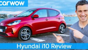 Imagen de un Hyundai i10 rojo aparcado frente al mar con un presentador sonriente señalándolo, logotipo de carwow en la esquina superior izquierda y banda inferior con el texto "Hyundai i10 Review", Ventajas y desventajas del Hyundai i10 2013: ¿vale la pena comprarlo?