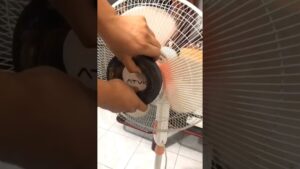 Manos desmontando la tapa central de un ventilador de pie blanco con rejilla metálica y aspas translúcidas sobre suelo de baldosas durante una reparación casera, descripción optimizada para accesibilidad y SEO que incluye la referencia Ventajas y desventajas de un motor DC de imán permanente esencial.