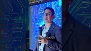 Ponente con gafas y chaqueta sostiene un micrófono de mano y un micrófono de solapa mientras habla frente a una pantalla azul con gráficos de circuito electrónico en una presentación sobre tecnología, Ventajas y desventajas de la inteligencia artificial: impacto real