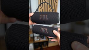 Fotografía de dos altavoces portátiles JBL negros con tejido texturizado y logo prominente, uno de mayor tamaño con asa y otro compacto sostenidos por manos delante de una estantería desenfocada, Jbl Charge 5 ventajas y desventajas: ¿vale la pena este altavoz?