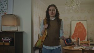 Mujer joven de pie en la entrada de una vivienda, con chaleco marrón sobre camisa blanca, bolso amarillo al hombro y sosteniendo un teléfono móvil cuya pantalla muestra una aplicación, en un salón con lámpara retro, mueble con una mini furgoneta de juguete, mesa con objetos y un cuadro naranja, imagen optimizada para SEO y accesibilidad que invita a la pregunta Ventajas y desventajas de Kayak app: ¿vale la pena usarla?