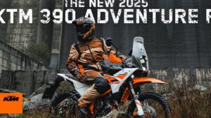 Motociclista con casco y traje naranja sentado en una KTM naranja y blanca de aventura con parabrisas alto, faro delantero y neumáticos de taco sobre terreno húmedo y vegetación frente a una estructura de hormigón industrial, perfecta imagen para ilustrar Ventajas y desventajas de la KTM 390 para elegir tu moto ideal.