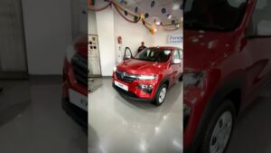 Vista frontal y lateral de un Renault Kwid rojo exhibido en un concesionario con la puerta abierta, decoraciones colgantes en el techo y un vendedor al fondo, optimizada para SEO y accesibilidad con la frase Renault Kwid 2025 ventajas y desventajas para elegir tu auto ideal.