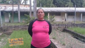 Mujer de pie con expresión seria, vistiendo camiseta rosa con mangas negras y las manos detrás de la espalda frente a camas de cultivo con plantaciones de cebollín y un cartel que dice Cebolleta, en un huerto rodeado por una valla metálica, palmas y un edificio deteriorado de fondo, Ventajas y desventajas de la UPETAI en Venezuela: impacto real